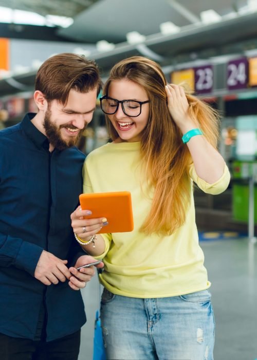 Portrait of couple standing in airport. She has long hair, glasses, sweater and jeans. He has beard, shirt, pants. They are looking on tablet.