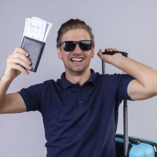 young handsome traveler man wearing black sunglasses holding air tickets standing with suitcase happy and exited smiling cheerfully over white background